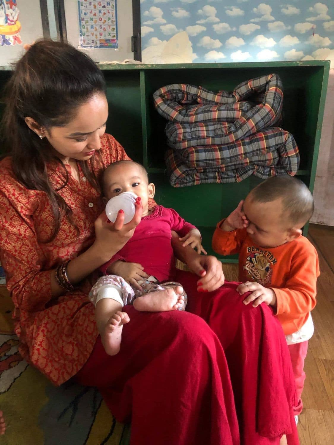 Pooja giving a baby a bottle in the Bal Mandir nursery while a toddler leans on her knee