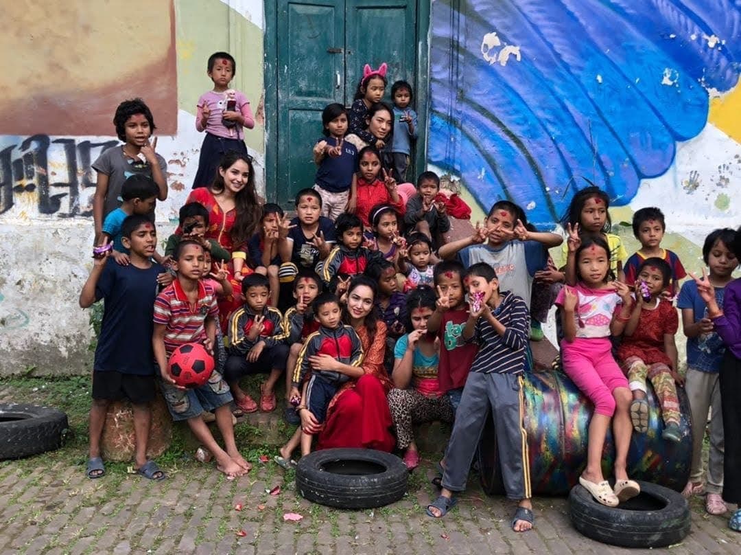 Pooja and a volunteer with a crowd of children in front of a painted wall at Bal Mandir