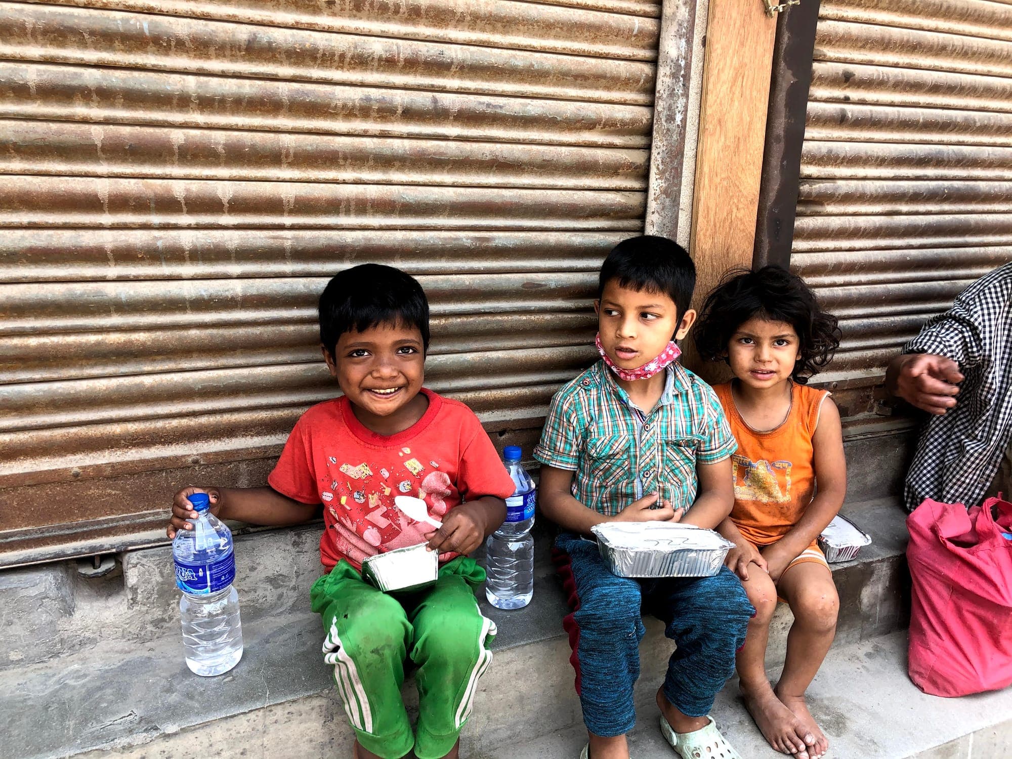 Three children sitting against a shutter with their meals and water