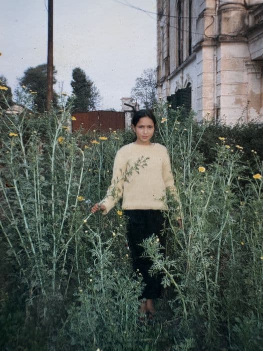 Pooja as a girl, standing in a field of tall yellow flowers beside an old building