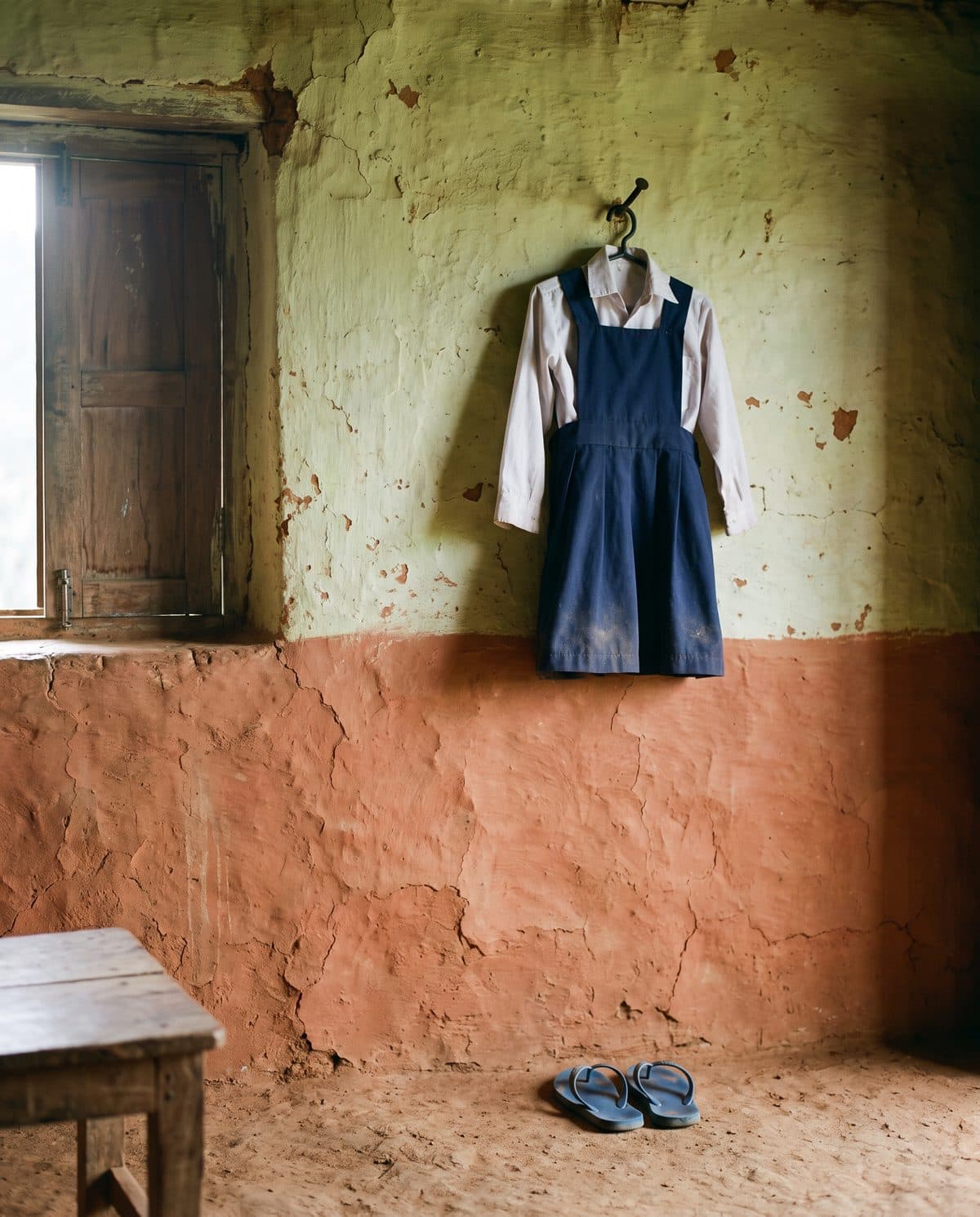 A child’s school uniform hanging unused on a nail in a village home, small flip-flops beneath it