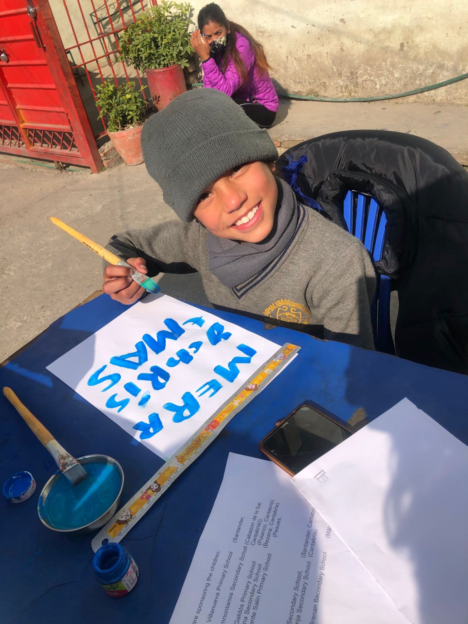 Nirjala smiling over a painted Christmas message, with a list of the Spanish schools that sponsor the children on the table beside it
