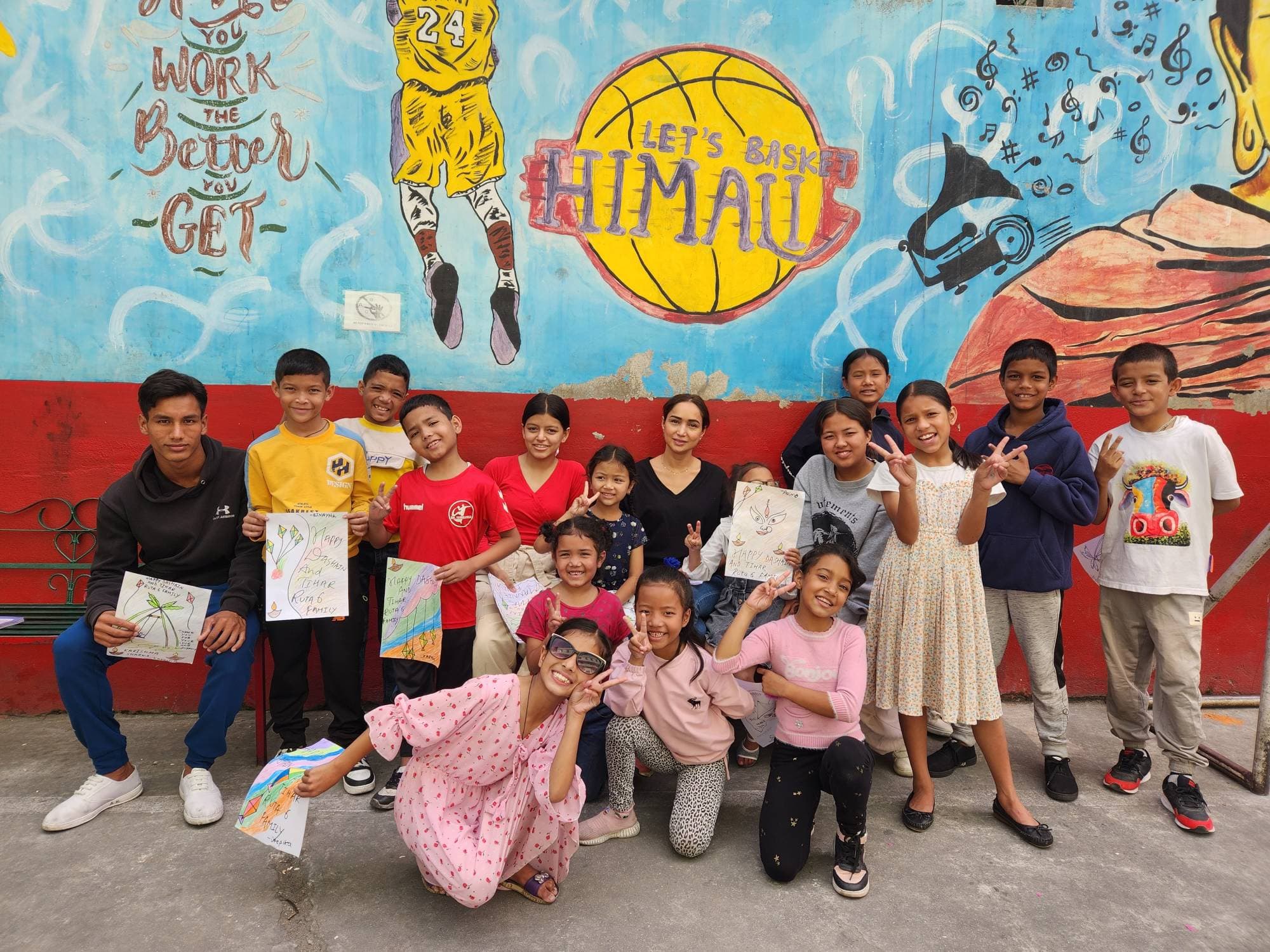 The children in front of a painted mural, holding up drawings that say Happy Dashain and Tihar, Ruta6 family