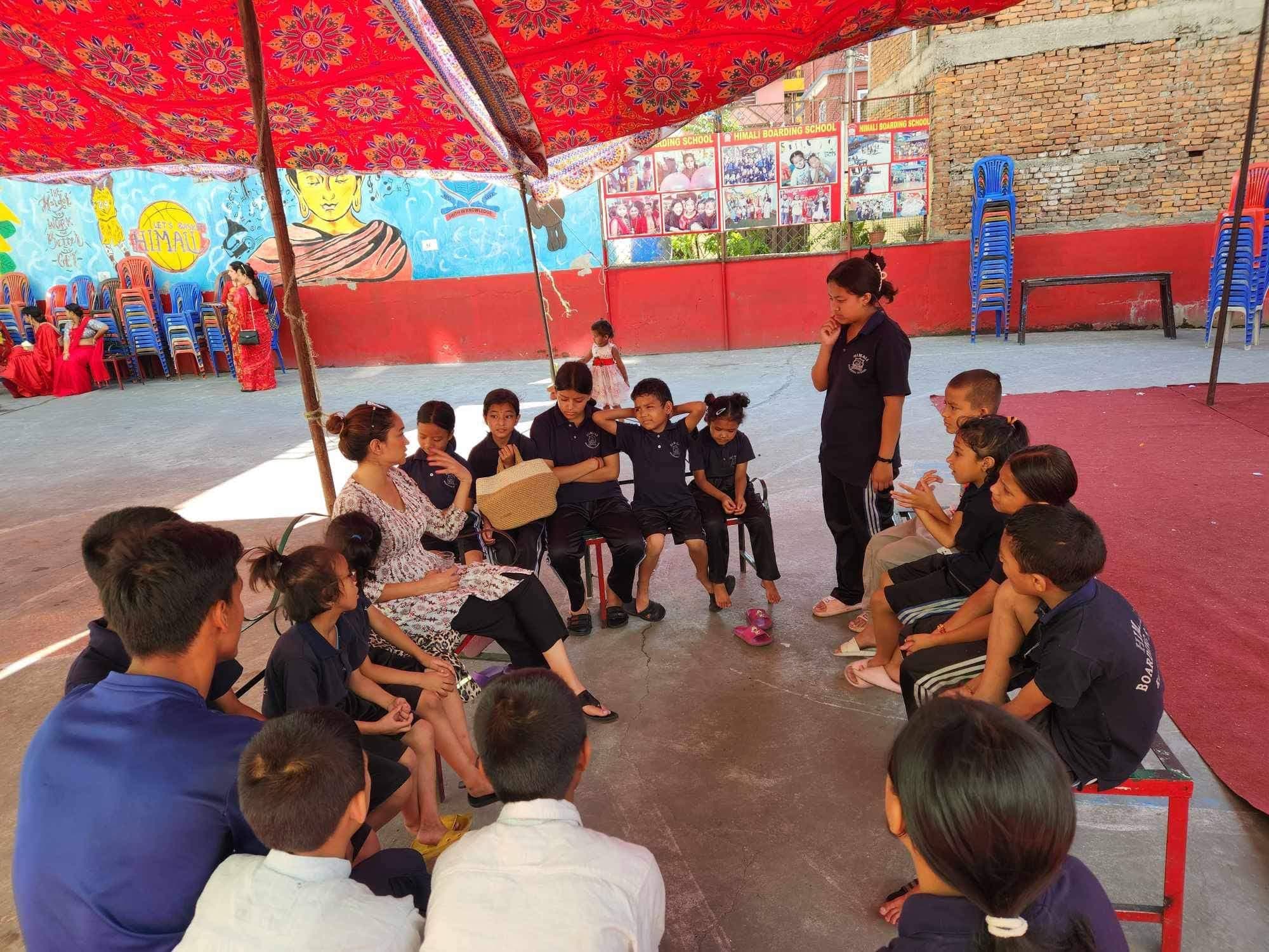 Pooja sitting in a circle of children in school uniform under a festival canopy, talking with them