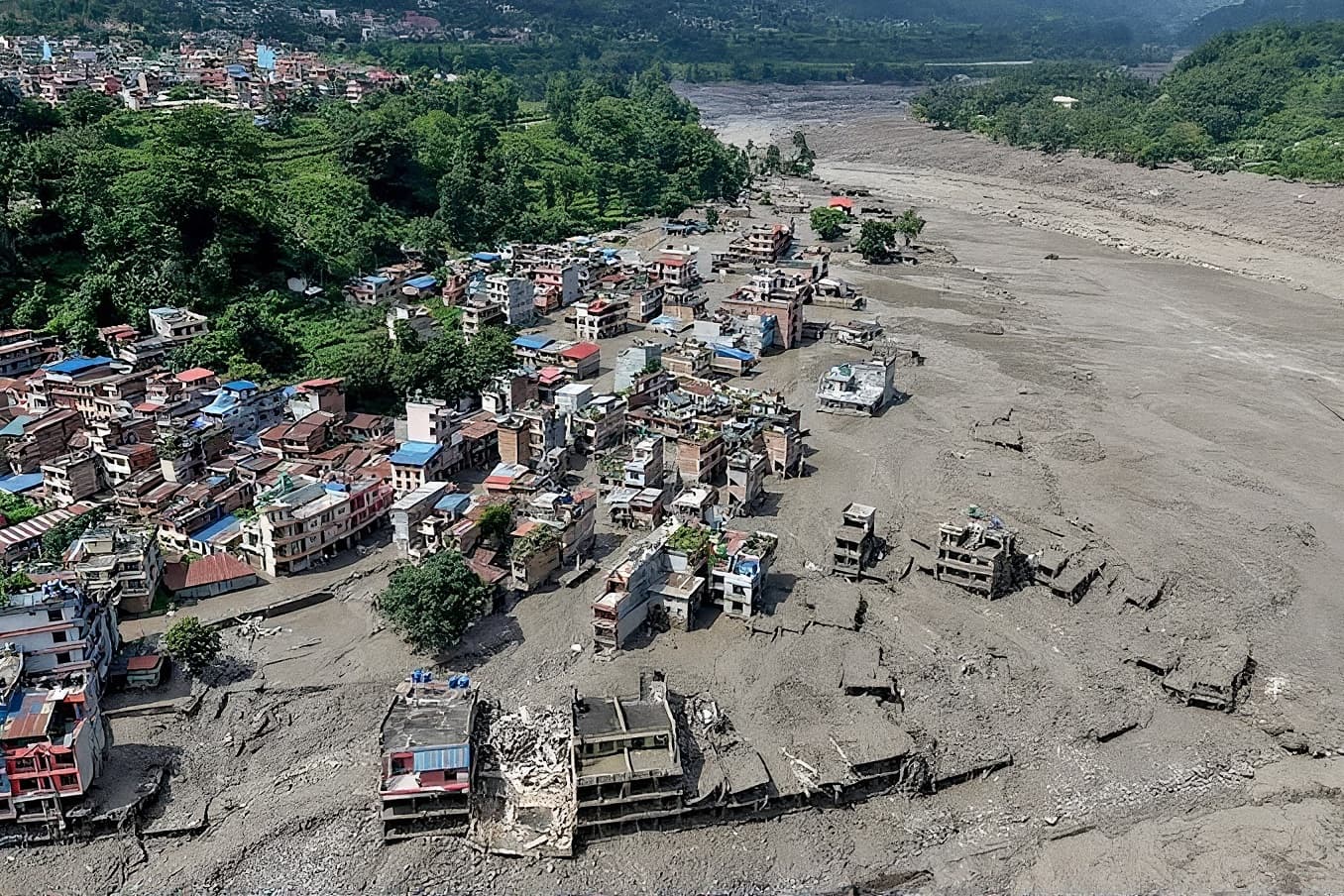 Aerial view of a riverside town half-buried in grey flood silt, houses standing in the mud