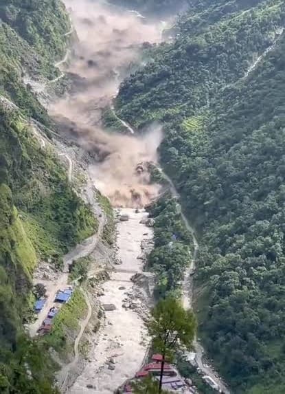A brown wall of floodwater and debris surging down a narrow forested gorge