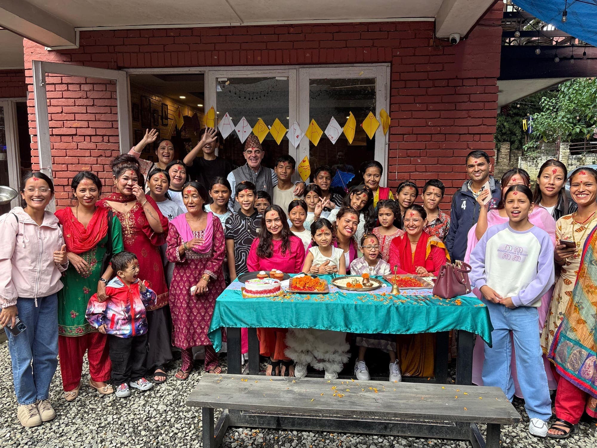 Teachers, families, Ruta6 and Pooja with the children around a table of flowers and cake, all wearing tika for Dashain