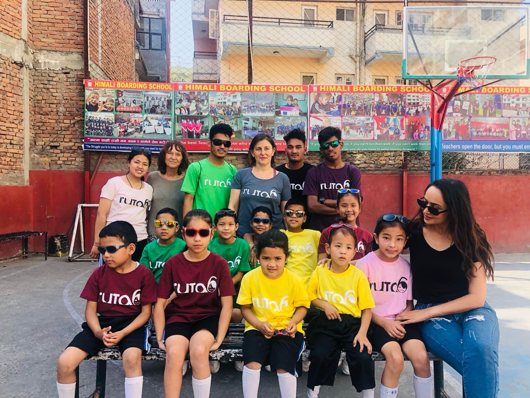 Ruta6 scholars in bright Ruta6 T-shirts and sunglasses on a bench at Himali Boarding School, with two Ruta6 volunteers and Pooja